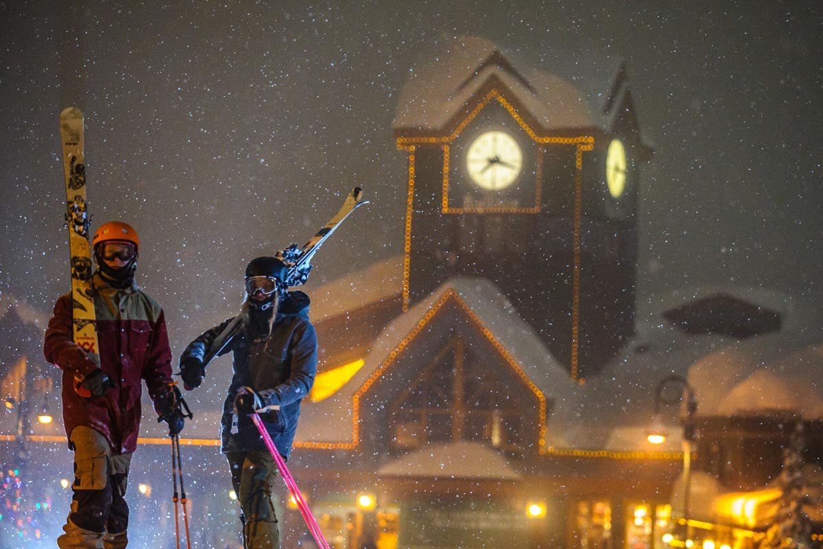 Two skiers walking through night snowfall at Big White, illuminated clock tower behind