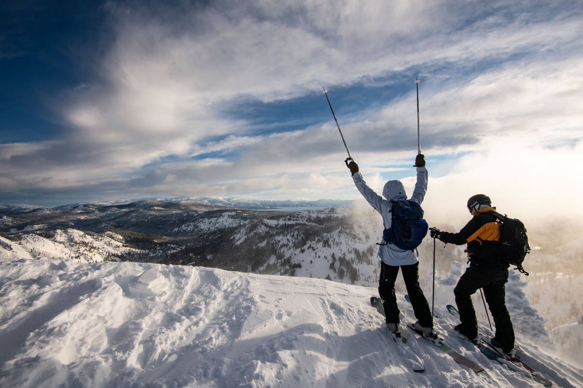 Two skiers celebrating on a snowy ridge above Lake Tahoe at Palisades Tahoe, California