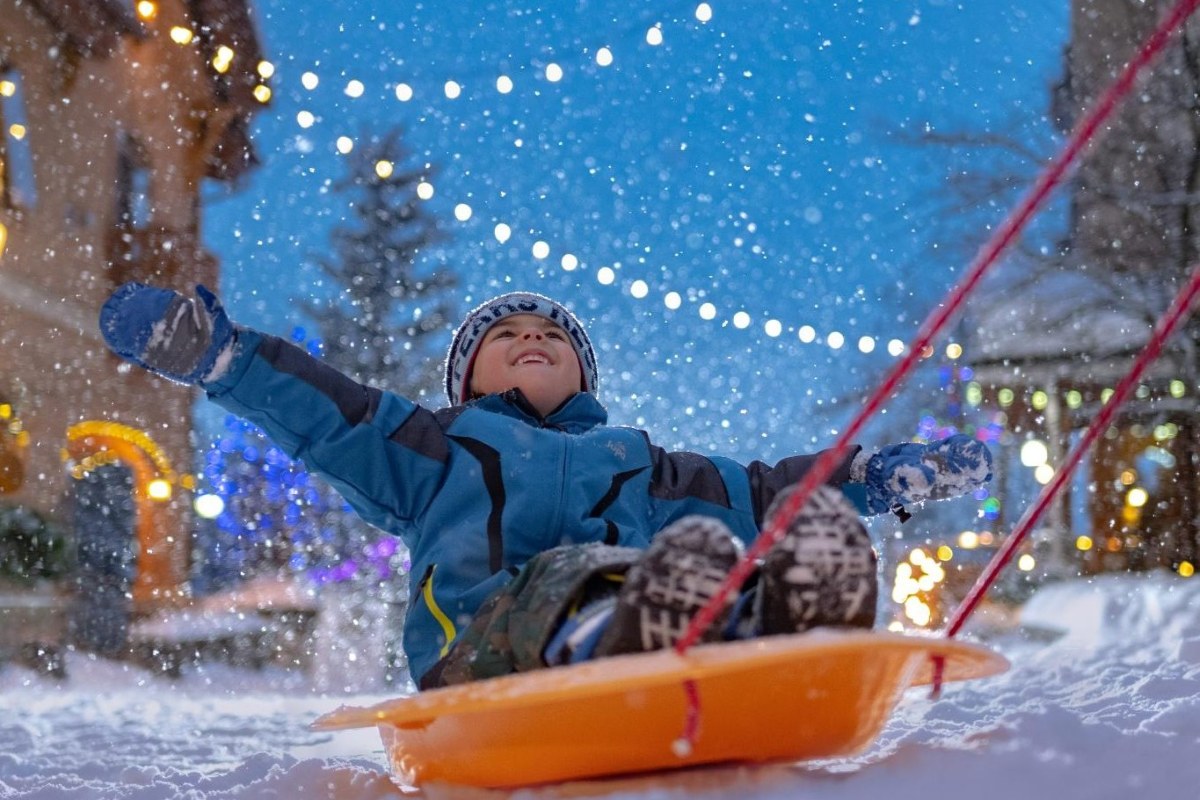 Child on a sled at Sun Peaks village during snowfall, string lights above